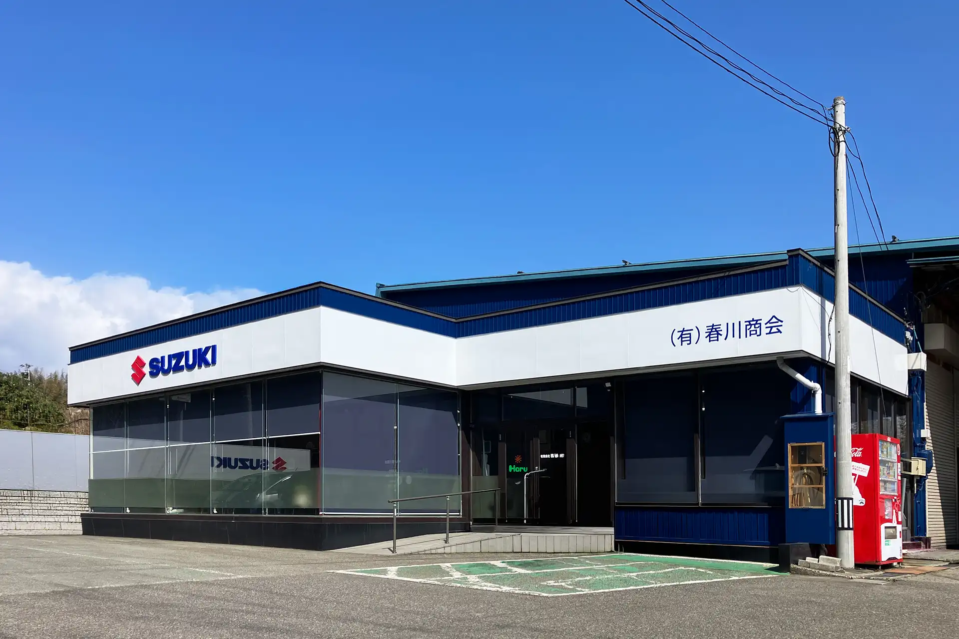 スズキのロゴと「(有)春川商会」の看板が掲げられた青と白の店舗外観